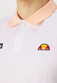 Gros plan sur un polo blanc avec un col rayé pêche, une patte de boutonnage et un logo en demi-cercle rouge et jaune sur la poitrine.