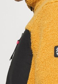 Giacca in pile con un corpo giallo testurizzato e maniche nere. Presenta una tasca con zip con dettagli rossi sul lato sinistro.
