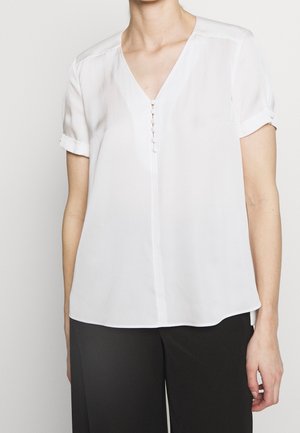 Kobieta ubrana w białą bluzkę z krótkim rękawem, dekoltem w kształcie litery V i guzikami obciągniętymi tkaniną, w połączeniu z czarnymi spodniami na jednolitym tle.