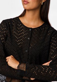 Blusa nera con un design a zigzag e dettagli perforati, scollo rotondo e chiusure a bottoni sul davanti e sui polsini.