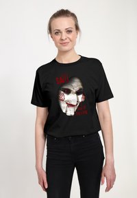 Černé bavlněné tričko s potiskem bílého obličeje loutky s červenými spirálkami, nápis „SAW Chci si zahrát hru“ červeně.