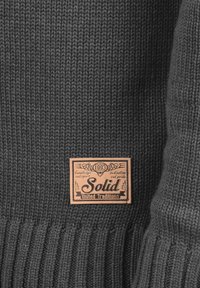 Dunkelgrauer Strickpullover-Stoff mit geripptem Saum. Verfügt über ein rechteckiges Lederetikett mit der eingeprägten Aufschrift "Solid United Traditions".
