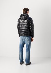 Man in een glanzende zwarte gewatteerde jas met kap, blauwe vervaagde jeans en zwarte schoenen, staande met zijn rug naar de camera op een plain witte achtergrond.
