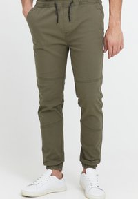 Olive green jogger pants with elastic waistband and drawstring, featuring side pockets and tapered cuffs. Paired with white sneakers.