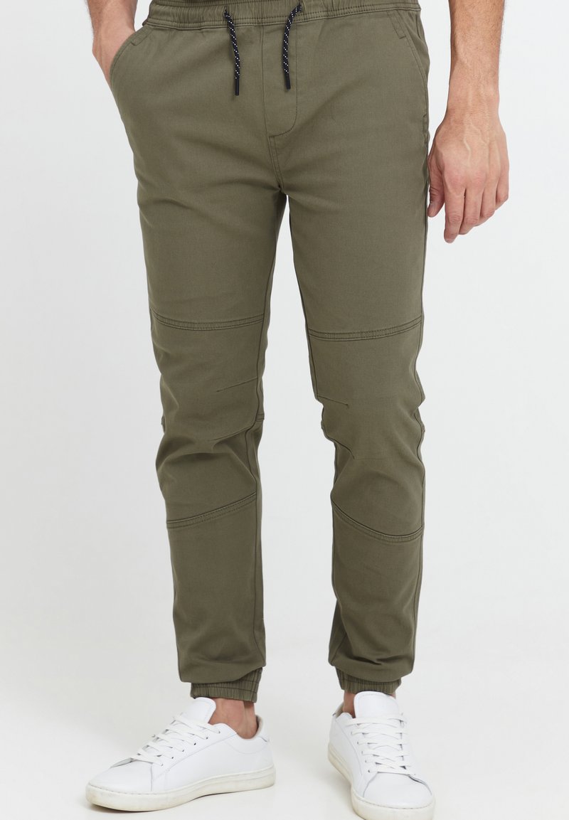 Olive green jogger pants with elastic waistband and drawstring, featuring side pockets and tapered cuffs. Paired with white sneakers.
