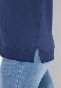 Felpa blu navy con orlo a coste, caratterizzata da uno spacco con bordi ondulati, abbinata a jeans azzurri. Tessuto texturizzato visibile.