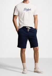 White cotton t-shirt with "Ralph" in blue script, paired with navy shorts featuring a drawstring. Styled with white sneakers and crew socks.