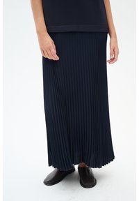 Granatowa plisowana spódnica z miękkiego materiału, z pionowymi fałdami, zwiewnym krojem, zestawiona z czarnymi butami na płaskiej podeszwie.