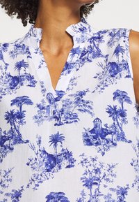 Hihaton valkoinen pusero, jossa sininen trooppinen maisemakuosi, palmupuita ja pieniä rakennuksia, sekä röyhelöillä koristeltu V-kaula-aukko.