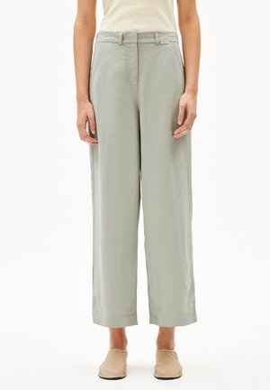 Personne portant un pantalon large gris clair avec poches latérales et des chaussures beige sans lacets, debout devant un fond blanc.