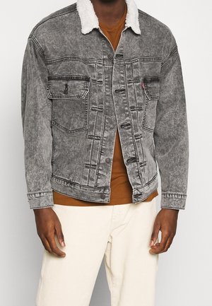 Personne portant une veste en jean grise avec un col en peau lainée blanc, par-dessus une chemise marron et un pantalon blanc, debout devant un fond uni.