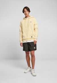Felpa gialla con tasca frontale e cordini, abbinata a pantaloni corti scuri con logo. Il modello indossa scarpe da ginnastica bianche. Sfondo neutro.