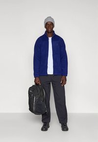 Mörkblå fleecetröja med dragkedja fram, bärs över en vit T-shirt, kombinerad med svarta byxor och en svart ryggsäck. Grå stickad mössa.