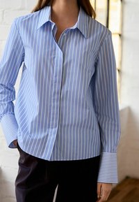 Chemise bleu clair à rayures blanches verticales, avec col pointu, manches longues et larges poignets détaillés. Coupe cintrée.