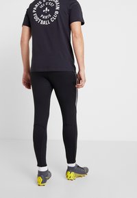 Zwarte sportlegging met een getextureerd zijpaneel, gecombineerd met een donker t-shirt met een wit logo. Grijze voetbalschoenen met gele accenten.