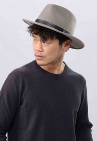 Homme aux cheveux courts et bouclés portant un chapeau fedora gris et un pull noir texturé, regardant vers sa gauche contre un fond clair.