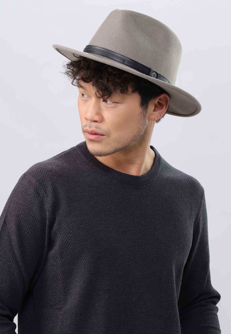 Homme aux cheveux courts et bouclés portant un chapeau fedora gris et un pull noir texturé, regardant vers sa gauche contre un fond clair.