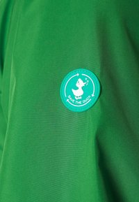 Tessuto della giacca verde con una texture liscia; presenta un logo circolare turchese "Salva l'anatra" con un'illustrazione di un'anatra bianca.