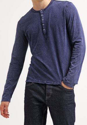 Granatowa koszula henley z długim rękawem, sześcioprzyciskowym obszyciem, miękką fakturą i subtelnymi detalami szwów. W połączeniu z ciemnymi dżinsami.