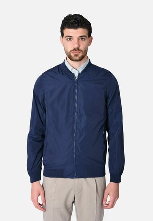 Uomo con giacca blu navy con zip sopra camicia con colletto e pantaloni beige, in piedi davanti a uno sfondo chiaro e uniforme.