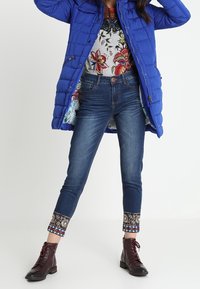 Manteau coupe-vent bleu, haut à imprimé floral, jean en denim foncé avec des poignets à motifs et bottines bordeaux, mettant en avant diverses textures et couleurs.