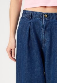 Pantalones de mezclilla en azul oscuro con pliegues en la cintura, que presentan un cierre de botón visible y una textura suave. Mano apoyada en la tela.