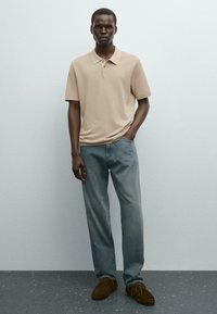 Beige polo shirt met korte mouwen, blauwe jeans en bruine suède schoenen. Model staat casual met de handen in de zakken tegen een neutrale achtergrond.
