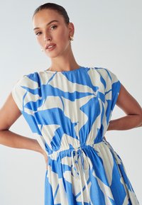 Sieviete ar saistītiem matiem, kas valkā zelta riņķa auskarus un zilu ar baltu abstraktu rakstu kleitu ar sašaurinātu vidukli, stāvot ar rokām uz gurniem.