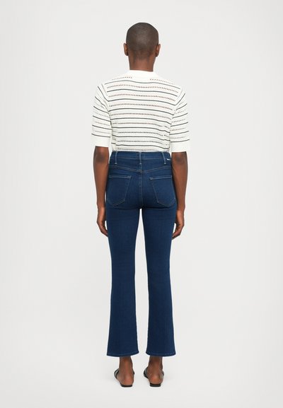 Een persoon staat met de rug naar de kijker en draagt een wit, kort mouwend gebreid top met horizontale strepen en donkerblauwe high-waisted flare jeans.