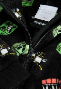 Mustavillainen fleece-takki, jossa on Minecraft-teemaisia kuvioita, mukaan lukien vihreä creeper, mustilla resoreilla ja vetoketjuyksityiskohdilla.