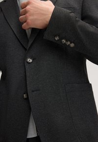 Dunkelgrauer Blazer aus strapazierfähigem Stoff, mit einer einzelnen Tasche, vier Knopf-Details an den Ärmeln und einem sichtbar hellgrauen Futter.