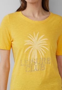 T-shirt jaune à manches courtes avec un col rond, mettant en avant un graphisme de palmier et le texte "LEISURE CLUB" dans une teinte légèrement plus sombre.