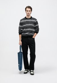 Black and gray striped sweater, black pants, black and white shoes, holding a blue denim tote bag with white text, on a neutral background.