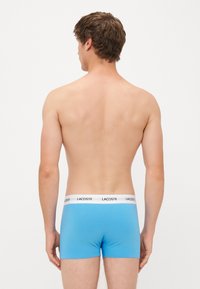 Homme de dos, portant un boxer bleu clair Lacoste avec une ceinture blanche, sur fond uni.