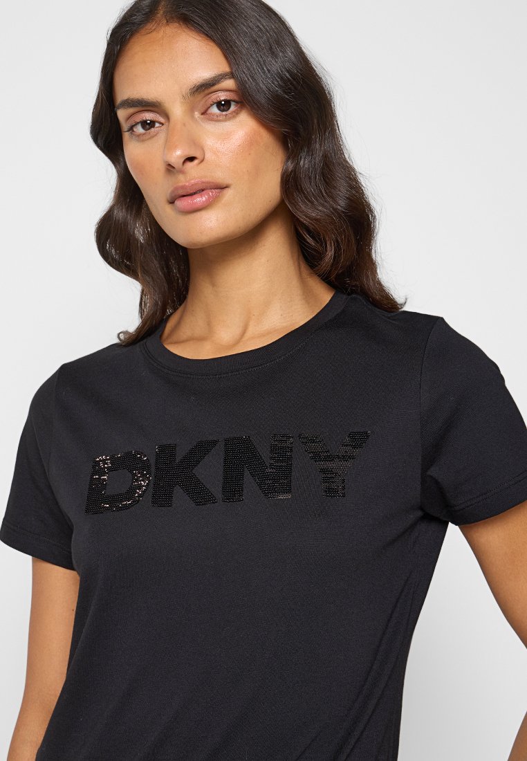 Camiseta negra de algodón con un logotipo texturizado de DKNY en lentejuelas. Mangas cortas, escote redondo y un diseño ajustado. Textura suave.