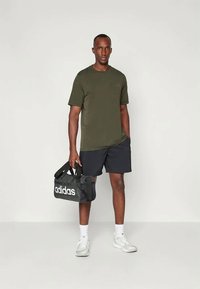 Camiseta verde de manga corta combinada con pantalones cortos negros, zapatillas blancas y un bolso de viaje negro de Adidas con el logo blanco, de pie sobre un fondo neutro.