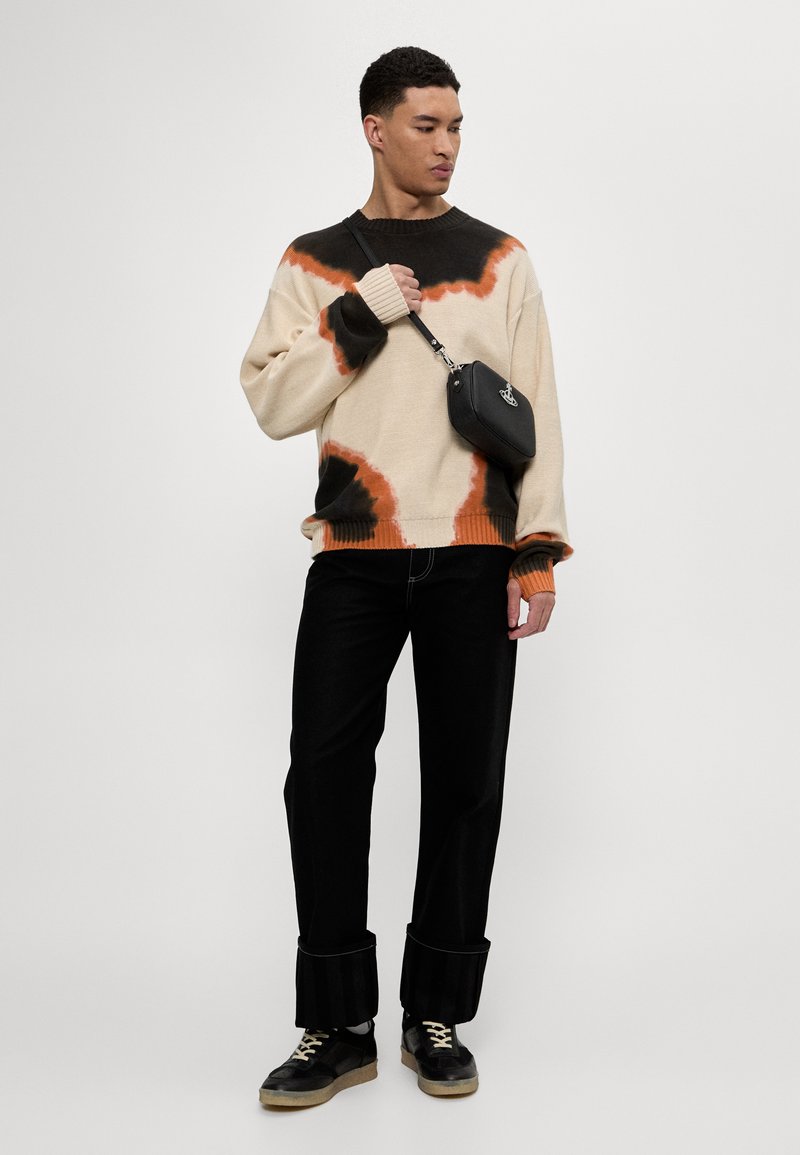 Beige gestrickter Pullover mit schwarzem und orangefarbenem Batikmuster, im Oversized-Schnitt. Kombiniert mit schwarzen Hosen mit ausgestellten Säumen und schwarzen Sneakers.