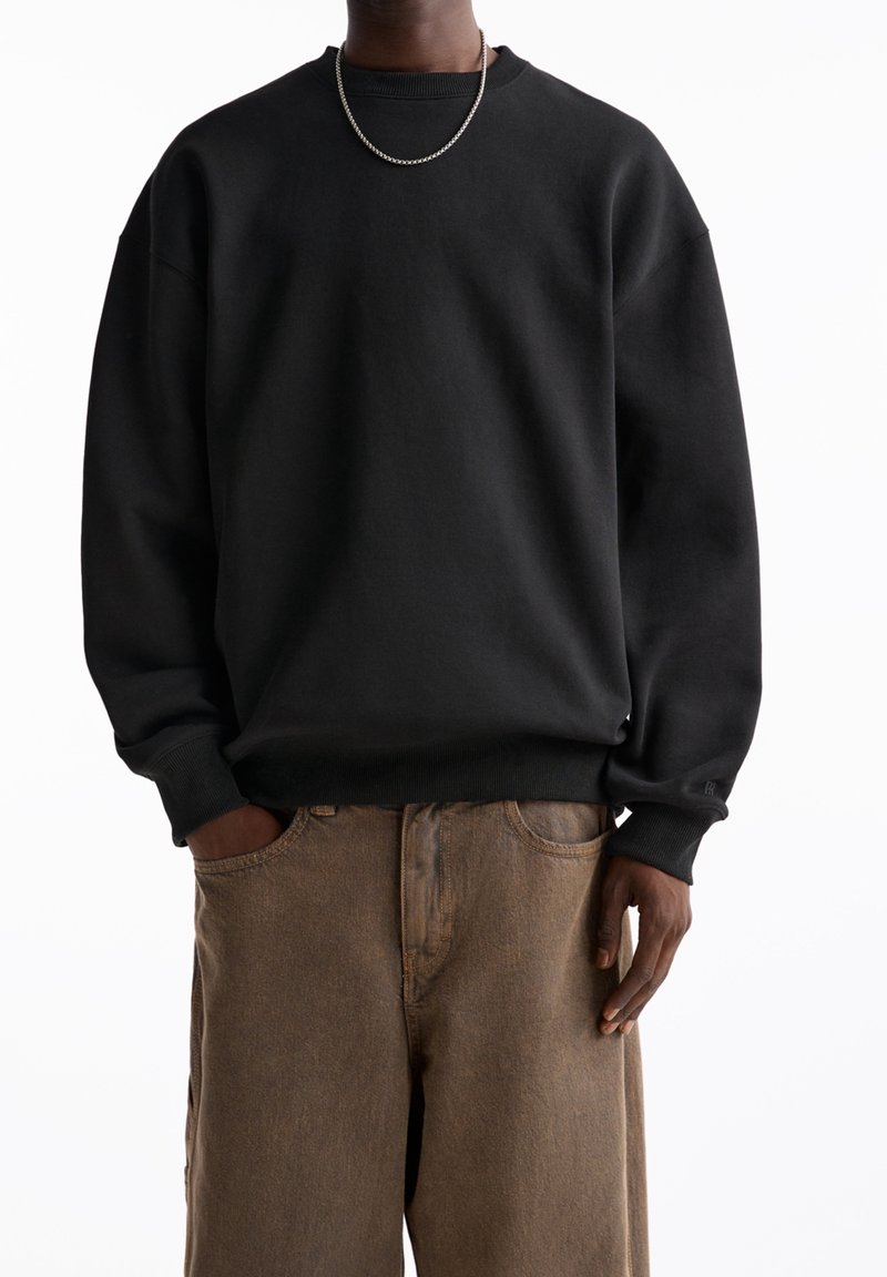 Personne portant un sweat-shirt noir à manches longues et un pantalon marron, une main dans la poche et un collier en chaîne argentée.