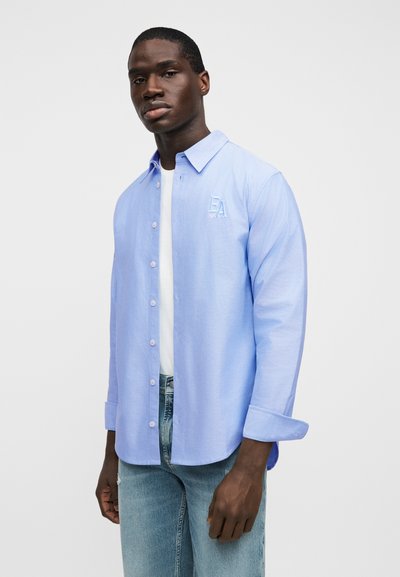 Το ανοιχτό μπλε πουκάμισο με κουμπιά διαθέτει γιακά, μακριά μανίκια και ένα διακριτικό κεντημένο λογότυπο "EA" στο στήθος. Φοριέται πάνω από ένα λευκό t-shirt.