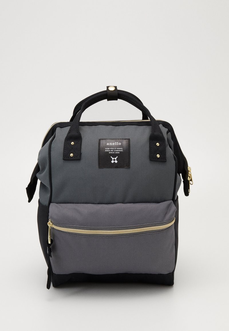 Kompaktowy, kwadratowy plecak z podwójnymi uchwytami u góry, przednią kieszenią na zamek oraz szaro-czarnym, kolorowym materiałem z naszywką z logo marki.