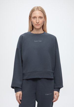 Kobieta o prostych blond włosach ubrana w ciemnoszarą bluzę Calvin Klein i dopasowane spodnie dresowe, stojąca na jasnym, jednolitym tle.