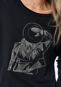 Zwarte lange mouwen shirt met witte borduursel van een persoon met lang haar die een hoed draagt, hand omhoog om de ogen te schaduw te geven, gedetailleerde texturen.