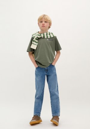 Grünes T-Shirt mit kurzen Ärmeln und Logo, blaue Denim-Jeans und braune Sneakers. Gestreifter grün-weißer Pullover um den Hals gebunden.
