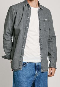 Graues Jeanshemd mit Button-Down-Kragen, langen Ärmeln, Brusttasche und kontrastierenden Nähten, getragen über einem weißen T-Shirt und blauen Jeans.