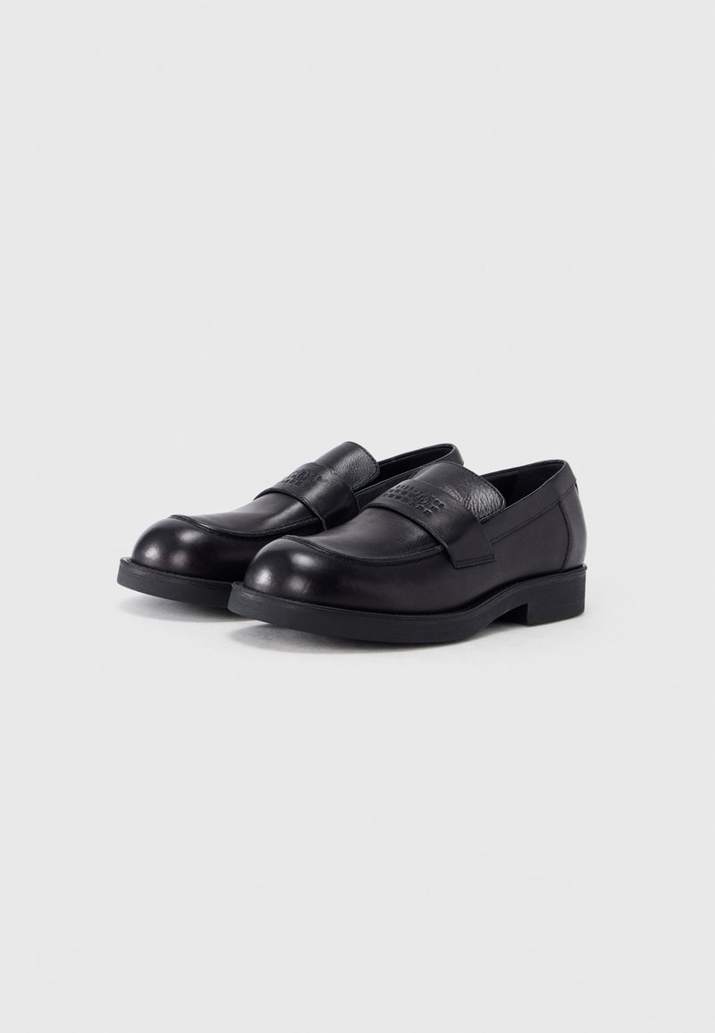 Par sorte læder slip-on loafers med runde tåspidser og en bred rem over vristen, vist på en ensartet lys baggrund.