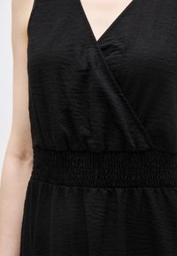 Μαύρο αμάνικο φόρεμα με κορσέ τύπου φιόγκος σε σχήμα V και ελαστική ζώνη στη μέση, φορεμένο από άτομο με γυμνά χέρια.