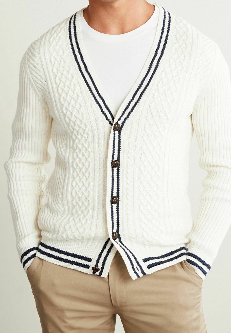 Cardigan en maille torsadée crème avec bordure rayée bleu marine, fermeture à boutons et texture côtelée. Associé à une chemise blanche et un pantalon beige.