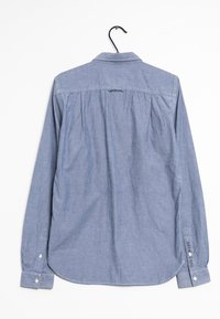 Vaaleansininen chambray-paita, jossa on pitkät hihat, nappikiinnitys hihansuissa, selässä laskokset ja kaareva helma. Takana on hienovarainen brändilappu.