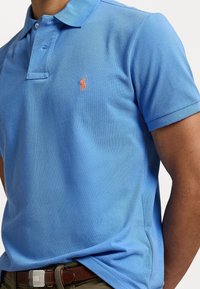 Man in een lichtblauw polo shirt met een oranje geborduurd logo, gestopt in een olijfgroene broek met een bruine riem.