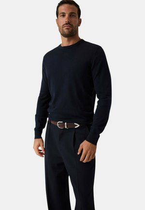 Homme portant un pull noir cintré à manches longues, un pantalon noir et une ceinture en cuir marron avec une boucle argentée, debout devant un fond blanc.